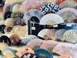 A display of various traditional Japanese folding fans, each with unique patterns and colors. Some fans feature floral designs, while others have geometric or abstract motifs. Price tags in yen are visible, indicating the fans are for sale.