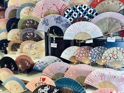 A display of various traditional Japanese folding fans, each with unique patterns and colors. Some fans feature floral designs, while others have geometric or abstract motifs. Price tags in yen are visible, indicating the fans are for sale.