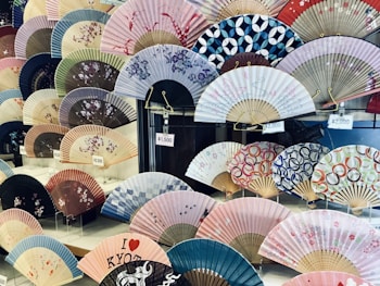 A display of various traditional Japanese folding fans, each with unique patterns and colors. Some fans feature floral designs, while others have geometric or abstract motifs. Price tags in yen are visible, indicating the fans are for sale.