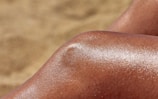 A close-up of a shiny, tanned knee against a blurred sandy background. The skin glistens, reflecting sunlight.