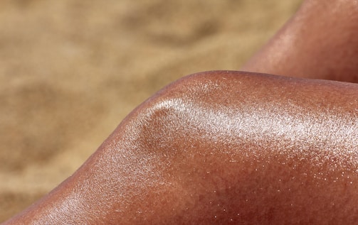 A close-up of a shiny, tanned knee against a blurred sandy background. The skin glistens, reflecting sunlight.