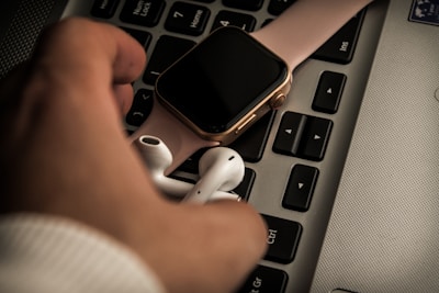 A close-up view of a smartwatch with a pink band placed on a laptop keyboard next to a pair of wireless earbuds. The hand of a person is partially visible in the frame, with fingers gently grasping the earbuds.