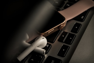 A smartwatch with a pink strap is placed on a laptop keyboard, accompanied by a pair of wireless earbuds. The scene is dimly lit, giving it a moody and tech-focused atmosphere.