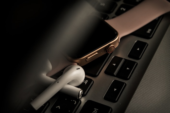 A smartwatch with a pink strap is placed on a laptop keyboard, accompanied by a pair of wireless earbuds. The scene is dimly lit, giving it a moody and tech-focused atmosphere.