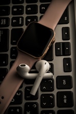 A smartwatch with a pink band and wireless earbuds are placed on top of a black computer keyboard.
