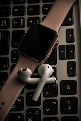 A smartwatch with a pink band and wireless earbuds are placed on top of a black computer keyboard.