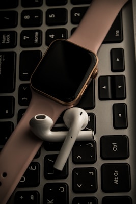 A smartwatch with a pink band and wireless earbuds are placed on top of a black computer keyboard.