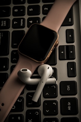 A smartwatch with a pink band and wireless earbuds are placed on top of a black computer keyboard.