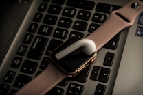 A pink smartwatch and a white wireless earbud are placed on a laptop keyboard. The devices are positioned diagonally across the keys, adding a modern and tech-oriented vibe to the composition.
