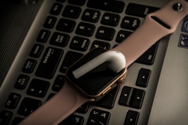 A pink smartwatch and a white wireless earbud are placed on a laptop keyboard. The devices are positioned diagonally across the keys, adding a modern and tech-oriented vibe to the composition.