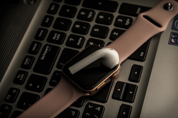 A pink smartwatch and a white wireless earbud are placed on a laptop keyboard. The devices are positioned diagonally across the keys, adding a modern and tech-oriented vibe to the composition.