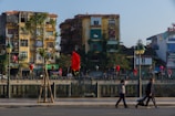 A vibrant urban scene with several old apartment buildings covered in greenery, featuring balconies and various advertisements. Red flags and lanterns are hung along the street, adding a festive or cultural ambiance. A few pedestrians walk along the foreground sidewalk, where a person is pushing a stroller.