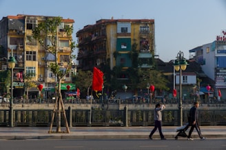 A vibrant urban scene with several old apartment buildings covered in greenery, featuring balconies and various advertisements. Red flags and lanterns are hung along the street, adding a festive or cultural ambiance. A few pedestrians walk along the foreground sidewalk, where a person is pushing a stroller.