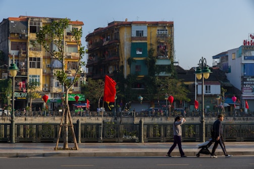 A vibrant urban scene with several old apartment buildings covered in greenery, featuring balconies and various advertisements. Red flags and lanterns are hung along the street, adding a festive or cultural ambiance. A few pedestrians walk along the foreground sidewalk, where a person is pushing a stroller.