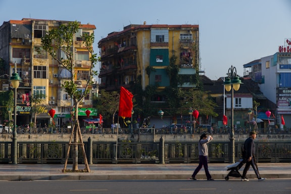 A vibrant urban scene with several old apartment buildings covered in greenery, featuring balconies and various advertisements. Red flags and lanterns are hung along the street, adding a festive or cultural ambiance. A few pedestrians walk along the foreground sidewalk, where a person is pushing a stroller.
