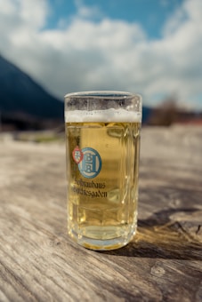 A clear glass mug filled with pale beer sits on a wooden surface, with a mountainous landscape blurred in the background. The mug has a logo and the text 'Hofbrauhaus Berchtesgaden' printed on it. The sky appears partly cloudy.
