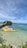A calm, clear sea stretches out under a bright blue sky, with inflatable banana boats in vibrant colors like yellow, orange, and blue floating near the shore. A lush, green island sits on the horizon in the background, with a small pier extending out to the water from the left side.