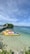 A calm, clear sea stretches out under a bright blue sky, with inflatable banana boats in vibrant colors like yellow, orange, and blue floating near the shore. A lush, green island sits on the horizon in the background, with a small pier extending out to the water from the left side.