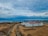 A large construction site with multiple white buildings featuring red roofs. The area is surrounded by barren land, and a visible pathway leads into the distance. The background showcases a wide expanse of water, likely a sea or ocean, with a cloudy sky overhead.