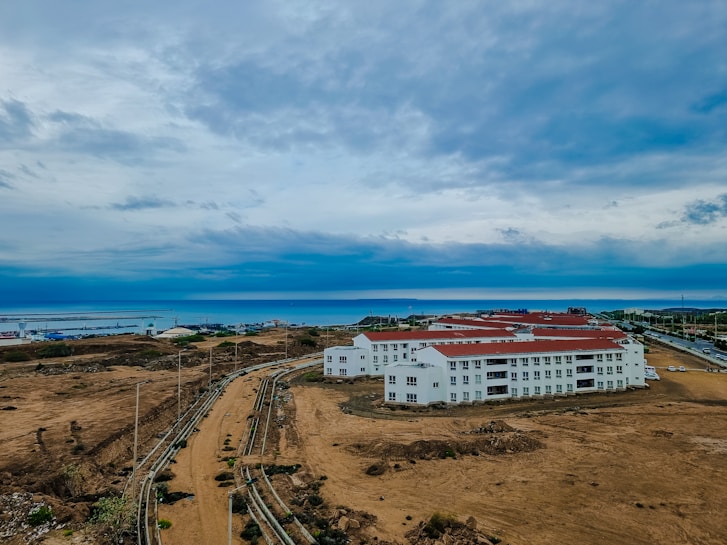 A large construction site with multiple white buildings featuring red roofs. The area is surrounded by barren land, and a visible pathway leads into the distance. The background showcases a wide expanse of water, likely a sea or ocean, with a cloudy sky overhead.