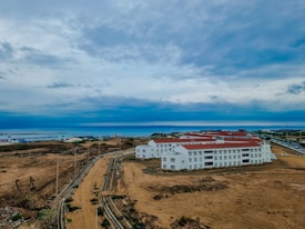 A large construction site with multiple white buildings featuring red roofs. The area is surrounded by barren land, and a visible pathway leads into the distance. The background showcases a wide expanse of water, likely a sea or ocean, with a cloudy sky overhead.