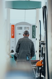 A person stands at a gas pump at a fuel station, viewed from behind. The person is wearing a jacket and appears to be refueling a vehicle. The gas pump shows numbers and has labels in what appears to be Arabic script. There are hoses and a bright orange handle visible on the pump.