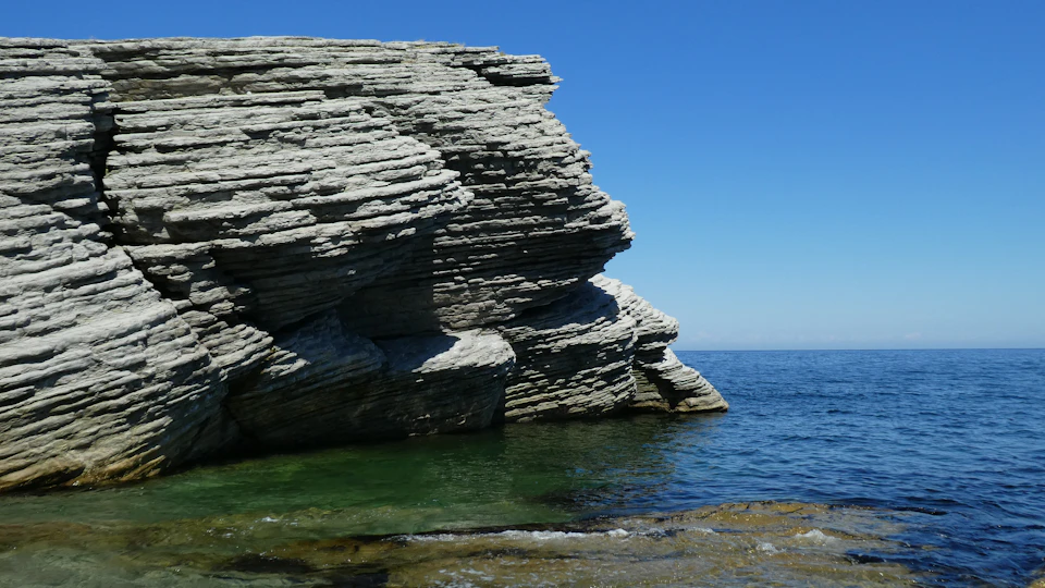 The image depicts a rocky cliff face with a large, jagged rock formation that appears to be made of gray and white stone. The cliff is situated on the edge of a body of water, which has a greenish hue. The sky above is clear blue, providing a beautiful backdrop for the rugged landscape below.
