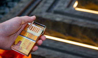 A hand is holding an open pack of cigarettes with a detailed illustration of a traditional building on the packaging. The pack features a warning label in Korean. In the background, there is a set of blurred stairs with yellow lighting.
