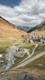 A small village nestled in a picturesque valley surrounded by brown and green hills with a winding river running through it. The village is composed of several buildings and scattered vehicles, with a dirt road curving alongside the river. Snow-capped mountains loom in the background under a partly cloudy sky.