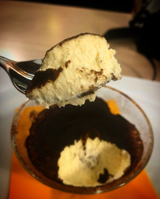A close-up of a spoon holding a portion of creamy dessert with a brown powder topping, held above a glass bowl containing the same dessert. The background is out of focus, highlighting the texture and colors of the dessert.