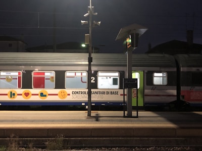 A train is stationed at a platform during nighttime. The train car is prominently marked with Italian writing related to basic health checks and free screenings. The platform is empty, with street lamps providing lighting, and the environment is dimly lit. Several windows on the train have visible signage indicating medical services.