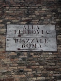 An old brick wall with a rectangular stone sign mounted on it. The sign has dark brown text and directional arrows pointing to the left, indicating 'Alla Ferrovia' on the top half and 'Piazzale Roma' on the bottom half.