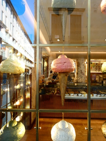 Large, artistic ice cream cone sculptures hang on display in a window, with a reflection of an old European street on the glass. Inside, the warm, inviting interior of a gelato shop is visible, showcasing a variety of flavors.