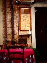 A rustic brick wall with a menu board displaying two Italian dishes with their prices. A wooden chair and a table with a red and white checkered tablecloth are in the foreground, suggesting an outdoor dining area. There are some potted plants and a dimly lit interior visible through a doorway.