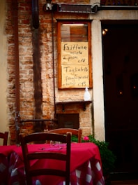 A rustic brick wall with a menu board displaying two Italian dishes with their prices. A wooden chair and a table with a red and white checkered tablecloth are in the foreground, suggesting an outdoor dining area. There are some potted plants and a dimly lit interior visible through a doorway.