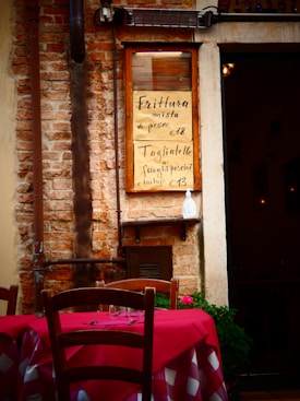 A rustic brick wall with a menu board displaying two Italian dishes with their prices. A wooden chair and a table with a red and white checkered tablecloth are in the foreground, suggesting an outdoor dining area. There are some potted plants and a dimly lit interior visible through a doorway.