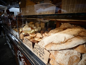 A variety of fresh bread and cheese are displayed in a glass case at a market stall. The assortment includes different shapes and sizes of loaves and rounds of cheese, some with labels in Italian. The interior of the market is dimly lit, with a focus on the textures and colors of the bread and cheese.