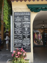 A street scene featuring a signboard listing various shops in an old arcade. The sign includes names and types of shops such as boutiques, gift shops, a sports shop, and a jeweller. The sign is attached to a building with green ivy growing on the side. In the background, people are seen walking past stores with colorful clothing displays. A potted plant with pink flowers is situated at the base of the sign.