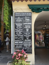 A street scene featuring a signboard listing various shops in an old arcade. The sign includes names and types of shops such as boutiques, gift shops, a sports shop, and a jeweller. The sign is attached to a building with green ivy growing on the side. In the background, people are seen walking past stores with colorful clothing displays. A potted plant with pink flowers is situated at the base of the sign.