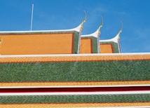 The image features a vibrant, traditional, Asian-style roof with overlapping tiles. The predominant colors are green and orange, and there are intricately designed golden spikes protruding from the top. The roof is set against a clear blue sky, suggesting a sunny day.