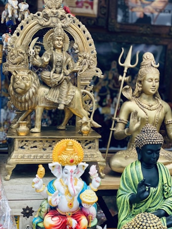 The image features an assortment of religious statues, including a golden deity riding a lion, a figure with a trident, a colorful elephant-headed figure, and a serene figure in a meditative pose. The vibrant and intricate designs display a range of expressions and symbolism.