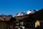 A picturesque mountain view with snow-capped peaks in the distance under a clear blue sky. In the foreground, there are several rustic, chalet-style buildings with sloped roofs, including one with a visible sign for 'Hotel Monzoni'. The surrounding area is lush with dense pine forests.
