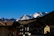 A picturesque mountain view with snow-capped peaks in the distance under a clear blue sky. In the foreground, there are several rustic, chalet-style buildings with sloped roofs, including one with a visible sign for 'Hotel Monzoni'. The surrounding area is lush with dense pine forests.