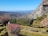 A scenic landscape featuring a structured garden with neatly trimmed hedges and colorful flowers. The garden is set on a hillside overlooking a vast expanse of rolling hills and distant mountains under a clear blue sky. A stone structure is visible on the edge of a rocky outcrop.