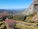 A scenic landscape featuring a structured garden with neatly trimmed hedges and colorful flowers. The garden is set on a hillside overlooking a vast expanse of rolling hills and distant mountains under a clear blue sky. A stone structure is visible on the edge of a rocky outcrop.