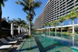 A modern, multi-story hotel with a curved facade overlooking a large swimming pool. Surrounding the pool are several palm trees and neatly arranged lounge chairs with closed umbrellas. The clear blue water of the pool reflects the greenery and the structure of the hotel. The sky is clear and blue.