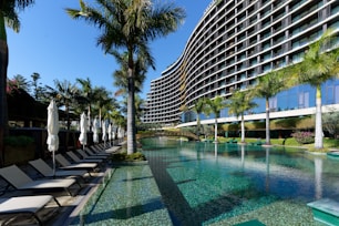 A modern, multi-story hotel with a curved facade overlooking a large swimming pool. Surrounding the pool are several palm trees and neatly arranged lounge chairs with closed umbrellas. The clear blue water of the pool reflects the greenery and the structure of the hotel. The sky is clear and blue.