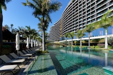 A modern, multi-story hotel with a curved facade overlooking a large swimming pool. Surrounding the pool are several palm trees and neatly arranged lounge chairs with closed umbrellas. The clear blue water of the pool reflects the greenery and the structure of the hotel. The sky is clear and blue.