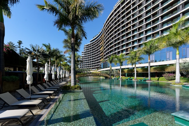 A modern, multi-story hotel with a curved facade overlooking a large swimming pool. Surrounding the pool are several palm trees and neatly arranged lounge chairs with closed umbrellas. The clear blue water of the pool reflects the greenery and the structure of the hotel. The sky is clear and blue.