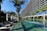 A modern, multi-story hotel with a curved facade overlooking a large swimming pool. Surrounding the pool are several palm trees and neatly arranged lounge chairs with closed umbrellas. The clear blue water of the pool reflects the greenery and the structure of the hotel. The sky is clear and blue.